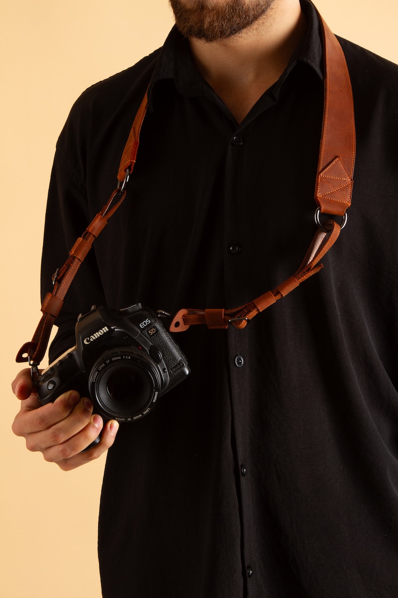 K&ouml;nnte beinhalten: Ein braunes Lederkameraband mit einem Metallring und einer daran befestigten Canon EOS 5D Kamera.