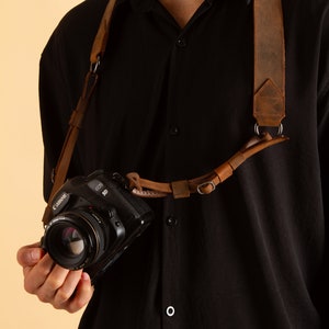 K&ouml;nnte beinhalten: Ein braunes Lederkameraband mit einer Metallschnalle und einer daran befestigten Canon EOS 5D Kamera.