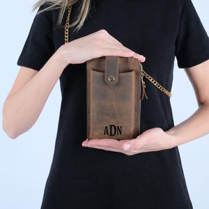 Pu&ograve; includere: Una borsa a tracolla per telefono in pelle marrone con una catena dorata. La borsa ha una chiusura a zip e una patta con chiusura a scatto. La borsa &egrave; personalizzata con le iniziali "ADN".