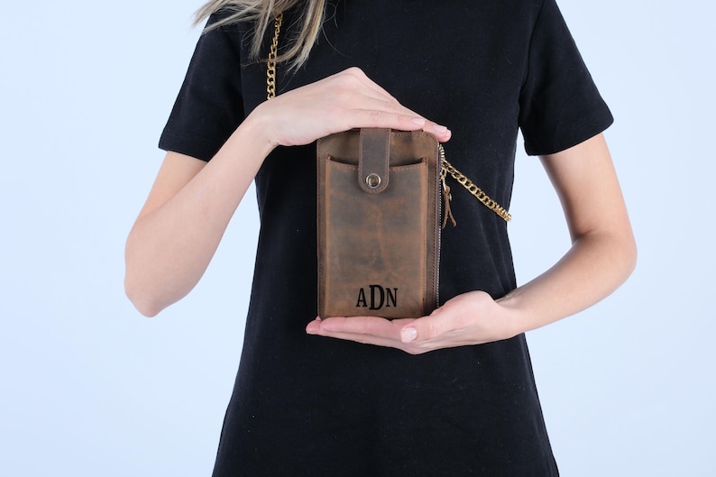 Pu&ograve; includere: Una borsa a tracolla per telefono in pelle marrone con una catena dorata. La borsa ha una chiusura a zip e una patta con chiusura a scatto. La borsa &egrave; personalizzata con le iniziali "ADN".