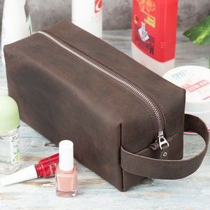 Pu&ograve; includere: Un beauty case in pelle marrone con chiusura a zip e manico in pelle. Il beauty case &egrave; appoggiato su una superficie in legno bianco con vari articoli da toeletta.