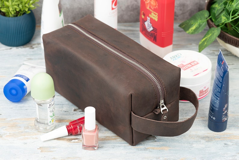 Pu&ograve; includere: Un beauty case in pelle marrone con chiusura a zip e manico in pelle. Il beauty case &egrave; appoggiato su una superficie in legno bianco con vari articoli da toeletta.