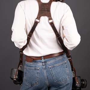 May include: A brown leather camera harness with the text "Zelary Photography" embossed on the back. The harness is worn over a white shirt and blue jeans, with two cameras attached. The leather is a rich, dark brown.