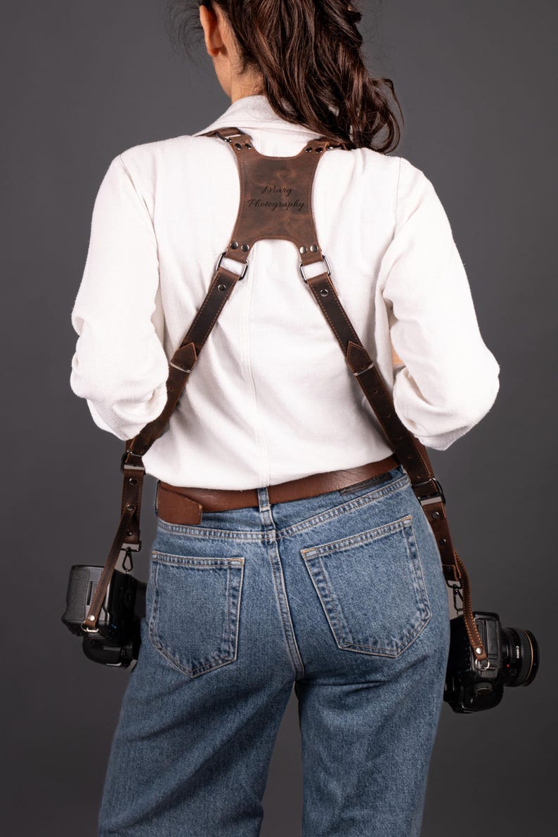 May include: A brown leather camera harness with the text "Zelary Photography" embossed on the back. The harness is worn over a white shirt and blue jeans, with two cameras attached. The leather is a rich, dark brown.