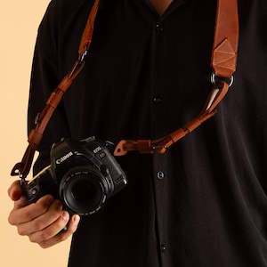 Pode incluir: Uma correia de c&acirc;mera de couro marrom com um anel de metal e uma c&acirc;mera Canon EOS 5D anexada.