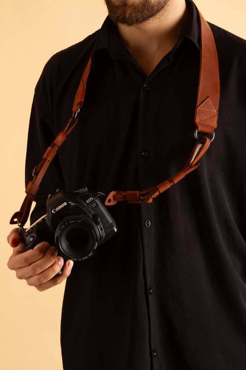 Pode incluir: Uma correia de c&acirc;mera de couro marrom com um anel de metal e uma c&acirc;mera Canon EOS 5D anexada.