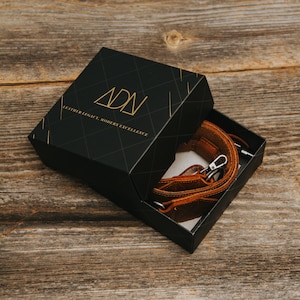 Pode incluir: Uma caixa preta com um logotipo dourado que diz "ADN Leather Legacy, Modern Excellence" est&aacute; aberta para revelar uma al&ccedil;a de c&acirc;mera de couro marrom.