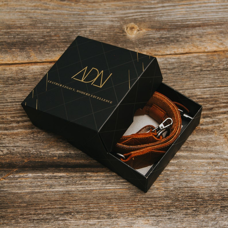 K&ouml;nnte beinhalten: Eine schwarze Box mit einem goldenen Logo, auf dem "ADN Leather Legacy, Modern Excellence" steht, ist ge&ouml;ffnet und zeigt ein braunes Lederkameraband.