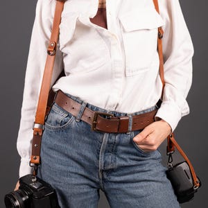 May include: A brown leather camera harness with a black camera attached. The harness is worn over a white shirt and blue jeans. A brown leather belt is also worn. The camera is hanging on the left side of the body.