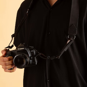 Pode incluir: Uma correia de c&acirc;mera preta com um anel de metal e uma c&acirc;mera preta com uma lente Canon EOS.