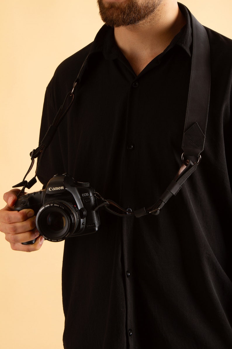 Pode incluir: Uma correia de c&acirc;mera preta com um anel de metal e uma c&acirc;mera preta com uma lente Canon EOS.
