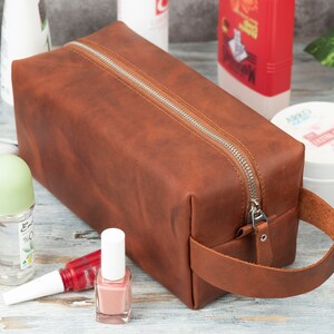 Pu&ograve; includere: Un beauty case da viaggio in pelle marrone con chiusura a zip e manico in pelle. La borsa &egrave; appoggiata su una superficie in legno con altri articoli da toeletta.