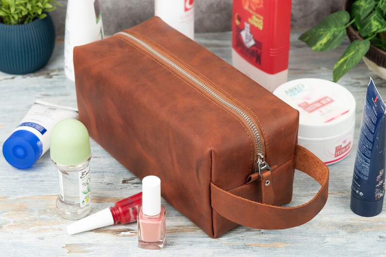 Pu&ograve; includere: Un beauty case da viaggio in pelle marrone con chiusura a zip e manico in pelle. La borsa &egrave; appoggiata su una superficie in legno con altri articoli da toeletta.