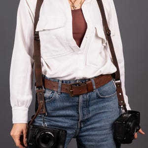 May include: A brown leather camera harness with two cameras attached. The harness is worn over a white shirt and blue jeans. The cameras are black and have lenses. A brown leather belt is also visible.