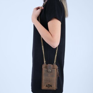 Pu&ograve; includere: Una borsa a tracolla per telefono in pelle marrone con una catena dorata. La borsa ha una chiusura a zip e una patta con chiusura a bottone. La borsa &egrave; personalizzata con le iniziali "ADN".