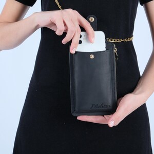 Pu&ograve; includere: Una custodia per telefono in pelle nera con una catena dorata e una cerniera. La custodia ha la scritta "Melissa" impressa sul davanti.