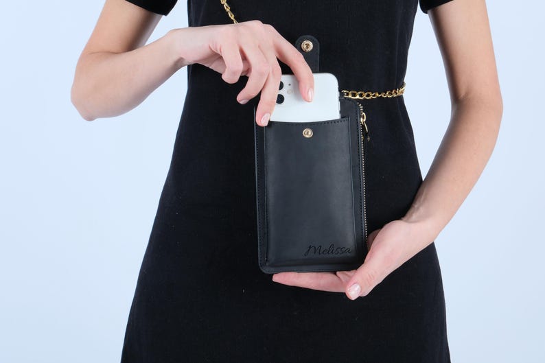 Pu&ograve; includere: Una custodia per telefono in pelle nera con una catena dorata e una cerniera. La custodia ha la scritta "Melissa" impressa sul davanti.