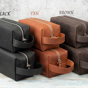 Pu&ograve; includere: Tre pile di pochette da toilette in pelle nera, marrone chiaro e marrone. Ogni pochette ha una chiusura a zip e un manico in pelle.