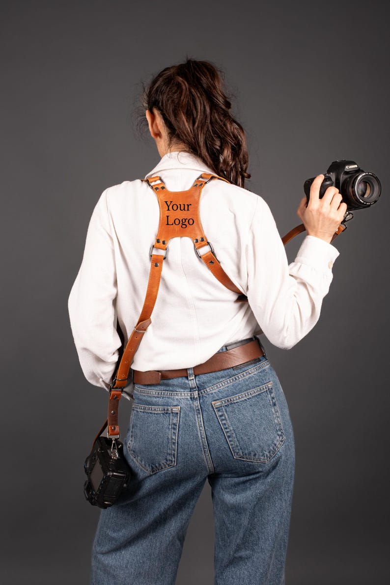 May include: A brown leather camera harness with the text "Your Logo" on the chest plate. The harness is worn over a white shirt and blue jeans. A black camera is attached to the harness.