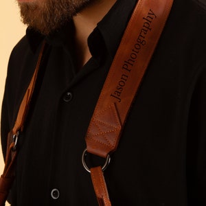 Pode incluir: Uma correia de c&acirc;mera de couro marrom com o texto "Jason Photography" gravado nela. A correia &eacute; usada sobre o ombro de uma pessoa que est&aacute; vestindo uma camisa preta.