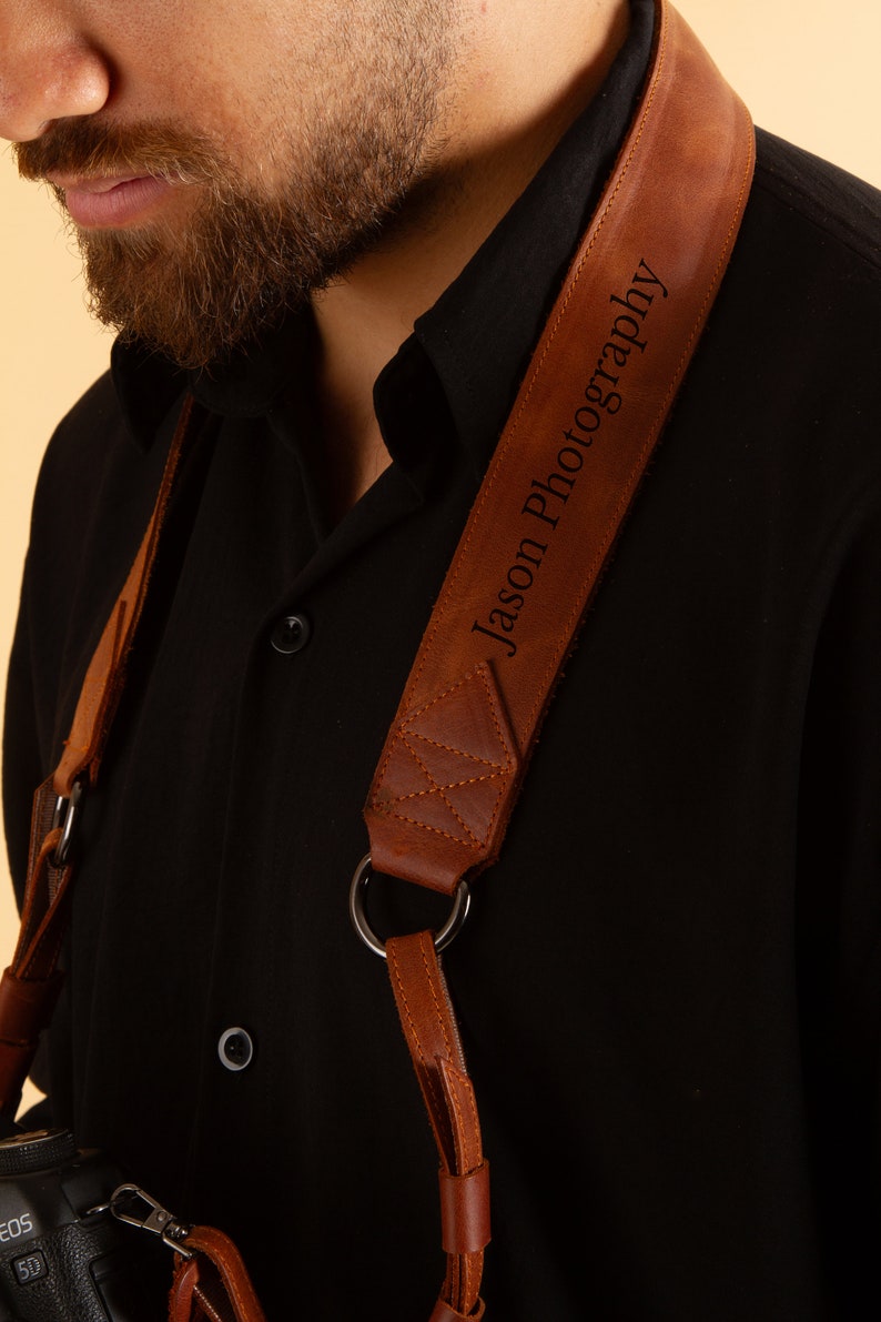 Pode incluir: Uma correia de c&acirc;mera de couro marrom com o texto "Jason Photography" gravado nela. A correia &eacute; usada sobre o ombro de uma pessoa que est&aacute; vestindo uma camisa preta.