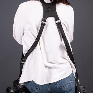 May include: Black leather camera harness with silver-tone fittings. The harness is embossed with the text "ADN MEDIA". Two black cameras are attached. The person is wearing a white shirt and blue jeans.