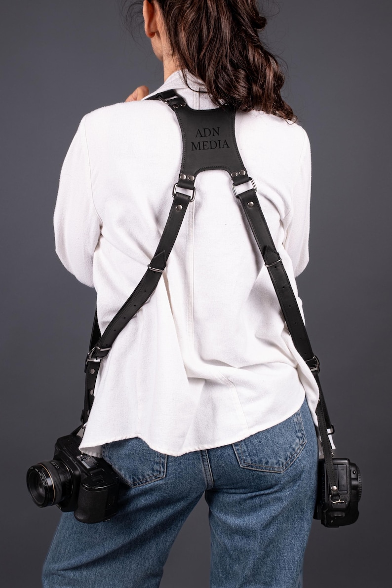 May include: Black leather camera harness with silver-tone fittings. The harness is embossed with the text "ADN MEDIA". Two black cameras are attached. The person is wearing a white shirt and blue jeans.