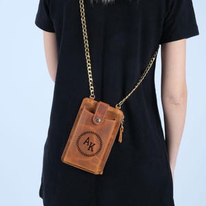 Pu&ograve; includere: Una borsa a tracolla per telefono in pelle marrone con una catena dorata. La borsa ha una chiusura a lembo con un bottone e una tasca con zip sul lato. La borsa &egrave; personalizzata con le iniziali "AK" in un cerchio con foglie.