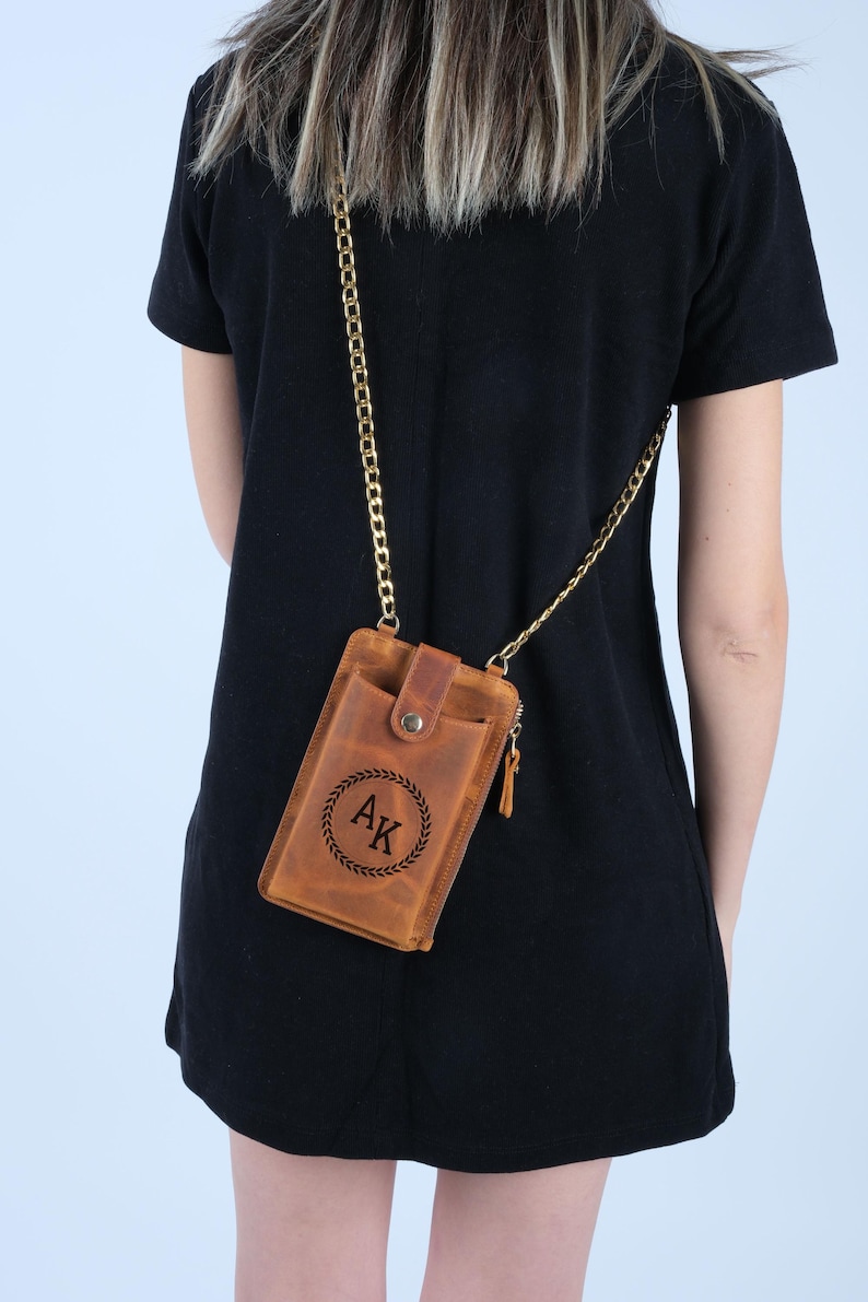 Pu&ograve; includere: Una borsa a tracolla per telefono in pelle marrone con una catena dorata. La borsa ha una chiusura a lembo con un bottone e una tasca con zip sul lato. La borsa &egrave; personalizzata con le iniziali "AK" in un cerchio con foglie.