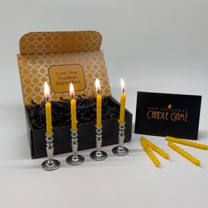 May include: A candle game set featuring four lit yellow candles in silver holders, a box with the text "Your New Tradition Starts Here", and a black card with "The Original Candle Game" printed on it. Additional yellow candles are also present.