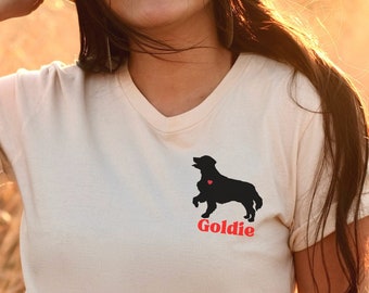 以下が含まれることがあります： クリーム色のTシャツを着た女性。黒い犬のシルエットと赤い文字で「Goldie」と書かれている。彼女は茶色の帽子をかぶり、長い茶色の髪をしている。