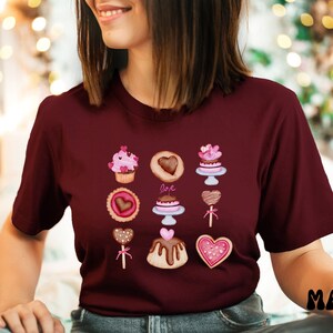 May include: A maroon t-shirt with a graphic design of various Valentine's Day themed desserts, including cupcakes, cookies, and cakes. The desserts are decorated with hearts and the word "Love" is featured on one of the cakes.