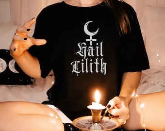Puede incluir: Camiseta negra con el texto "Hail Lilith" en fuente gótica, sobre un símbolo de luna creciente y cruz. La imagen incluye una vela encendida en un soporte y una almohada con fases lunares.