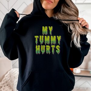 Puede incluir: Sudadera con capucha negra con el texto "MY TUMMY HURTS" en letras verdes que gotean.