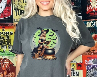 Może przedstawiać: Szary t-shirt z nadrukiem czarownicy trzymającej kociołek. Czarownica ma na sobie czarną sukienkę i spiczasty kapelusz. Jest otoczona nietoperzami i pełnią.