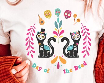 Könnte beinhalten: Weißes T-Shirt mit einem Día de Muertos-Motiv mit zwei schwarzen Katzen mit Zucker Totenköpfen, Blumen und anderen festlichen Elementen. Der Text "Day of the Dead" ist unter den Katzen gedruckt.