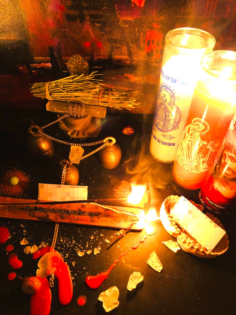 May include: A dark surface with a lit incense stick, two burning candles, and a bundle of dried herbs. The candles are labelled "Our Lady of Suyapa" and "Gran Poder". There are also several small bells, a chain, and a small piece of paper.