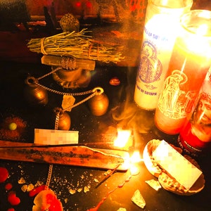 May include: A dark surface with a lit incense stick, two burning candles, and a bundle of dried herbs. The candles are labeled "Our Lady of Suyapa" and "Gran Poder". There are also several small bells, a chain, and a small piece of paper.