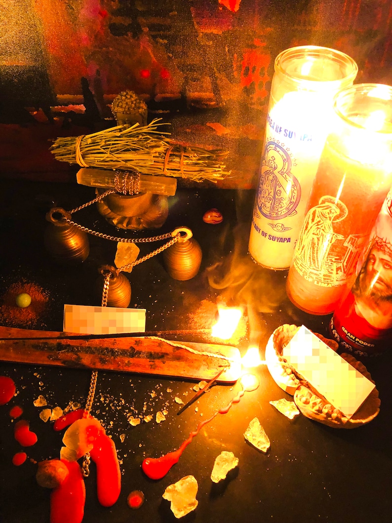 May include: A dark surface with a lit incense stick, two burning candles, and a bundle of dried herbs. The candles are labeled "Our Lady of Suyapa" and "Gran Poder". There are also several small bells, a chain, and a small piece of paper.