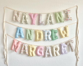 Könnte beinhalten: Ein dreistufiges Stoffbuchstaben-Banner mit den Namen NAYLANI, ANDREW und MARGARET. Jeder Buchstabe ist einzeln gefüllt und aus verschiedenen pastellfarbenen Stoffen gefertigt, darunter Blumenmuster und Vichy-Karos, aufgehängt an weißen Bändern.
