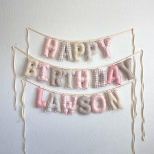 K&ouml;nnte beinhalten: Ein dreistufiges Stoffbanner mit der Aufschrift HAPPY BIRTHDAY LAWSON. Die ausgestopften Buchstaben bestehen aus einer Mischung aus einfarbigen rosa, beigen und wei&szlig;en Stoffen mit Karomustern, Streifen und Punkten, befestigt an cremefarbenen B&auml;ndern.