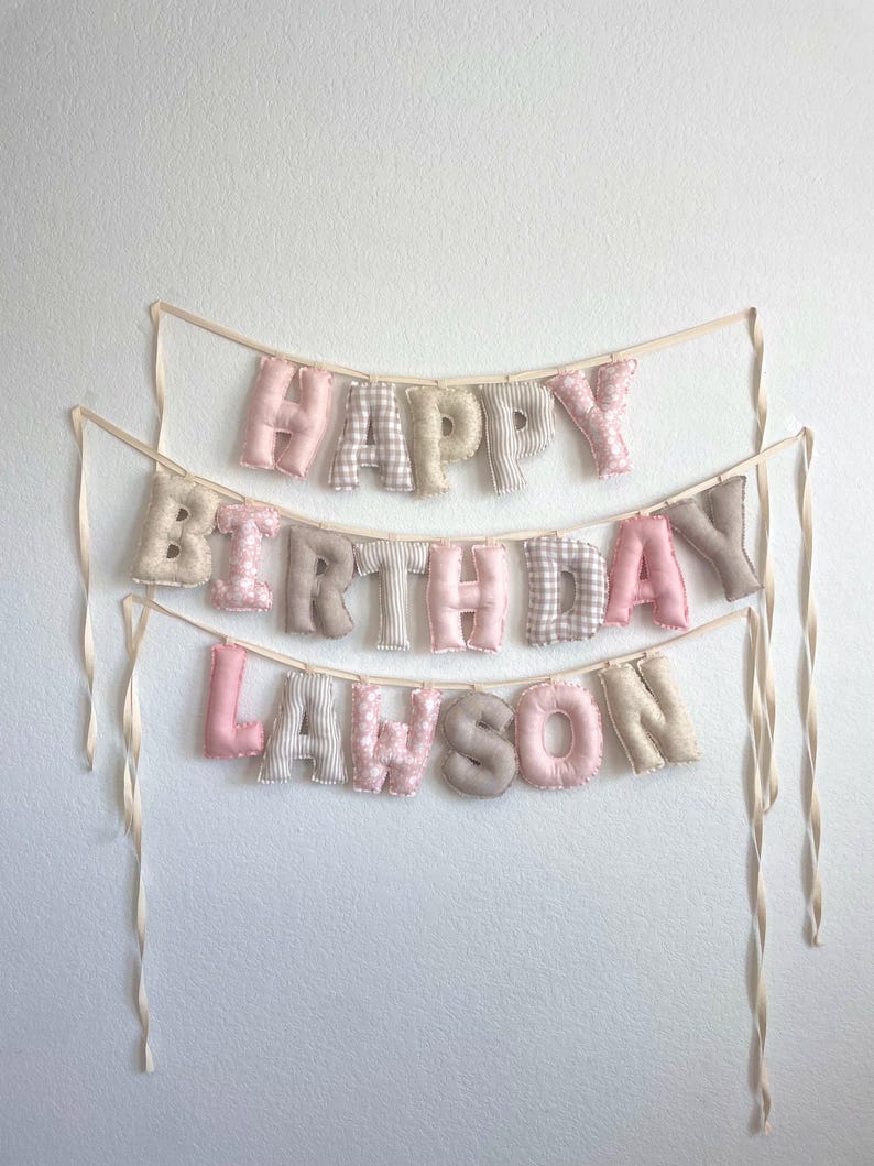 K&ouml;nnte beinhalten: Ein dreistufiges Stoffbanner mit der Aufschrift HAPPY BIRTHDAY LAWSON. Die ausgestopften Buchstaben bestehen aus einer Mischung aus einfarbigen rosa, beigen und wei&szlig;en Stoffen mit Karomustern, Streifen und Punkten, befestigt an cremefarbenen B&auml;ndern.