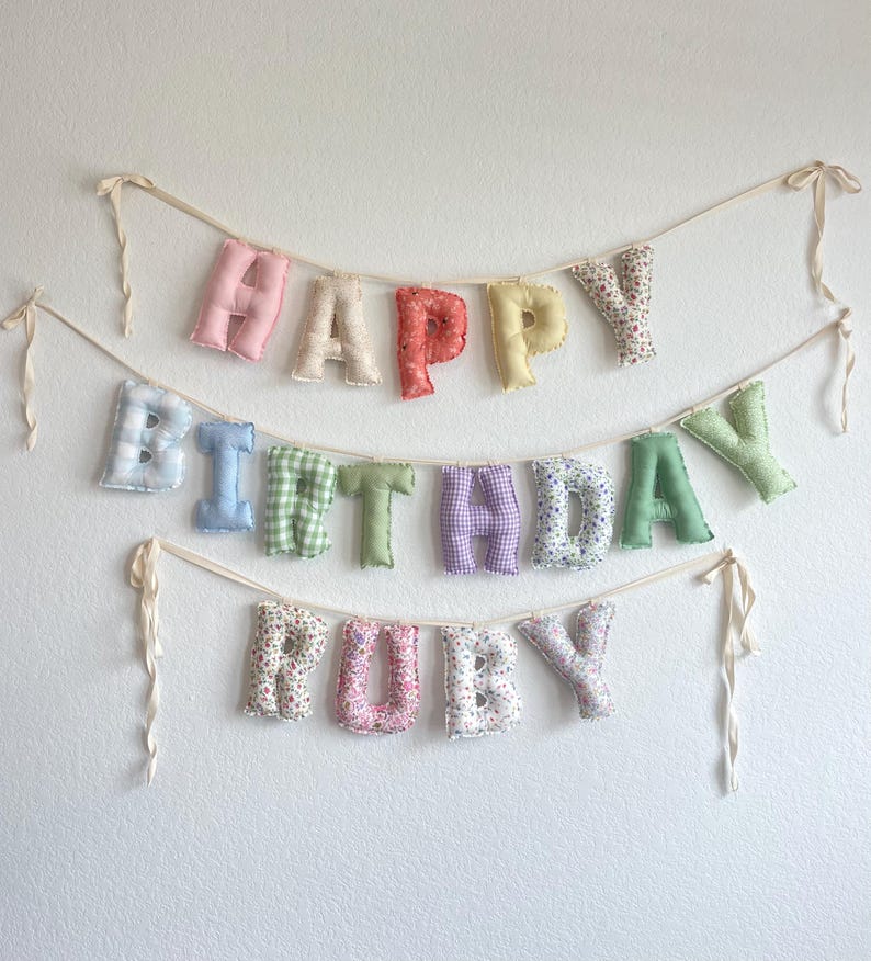 K&ouml;nnte beinhalten: Ein dreireihiges Stoffbanner mit der Aufschrift HAPPY BIRTHDAY RUBY. Die ausgestopften, weichen Buchstaben zeigen verschiedene Muster wie Vichy-Karos, Blumen und einfarbige Pastellt&ouml;ne, die auf einem cremefarbenen Band aufgereiht sind.