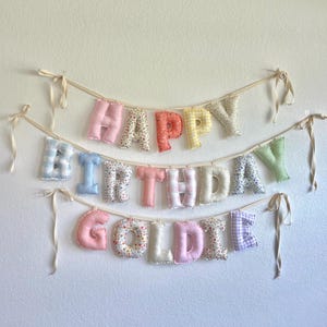 K&ouml;nnte beinhalten: Ein Stoffbanner mit drei Reihen aus gepolsterten, weichen Buchstaben, die HAPPY BIRTHDAY GOLDIE bilden. Jeder Buchstabe ist aus verschiedenen pastellfarbenen Stoffen mit Blumen-, Vichy- und Streifenmustern gefertigt und an cremefarbenen B&auml;ndern befestigt.