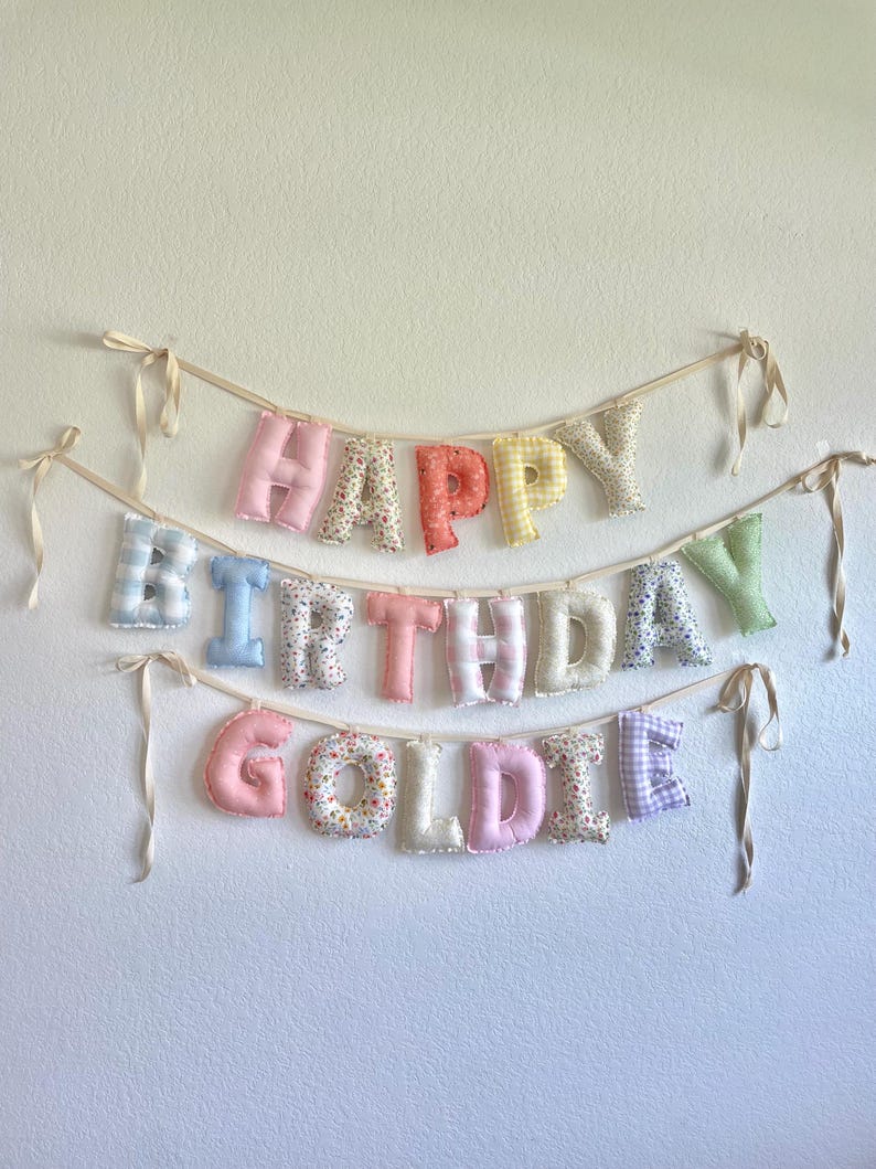 K&ouml;nnte beinhalten: Ein Stoffbanner mit drei Reihen aus gepolsterten, weichen Buchstaben, die HAPPY BIRTHDAY GOLDIE bilden. Jeder Buchstabe ist aus verschiedenen pastellfarbenen Stoffen mit Blumen-, Vichy- und Streifenmustern gefertigt und an cremefarbenen B&auml;ndern befestigt.