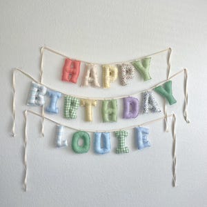K&ouml;nnte beinhalten: Ein Stoffbanner mit ausgestopften, weichen Buchstaben, die HAPPY BIRTHDAY LOUIE buchstabieren. Die Buchstaben sind aus verschiedenen pastellfarbenen Stoffen mit Vichy-Karo-, Streifen- und Blumenmustern gefertigt und an d&uuml;nnen, cremefarbenen B&auml;ndern befestigt.