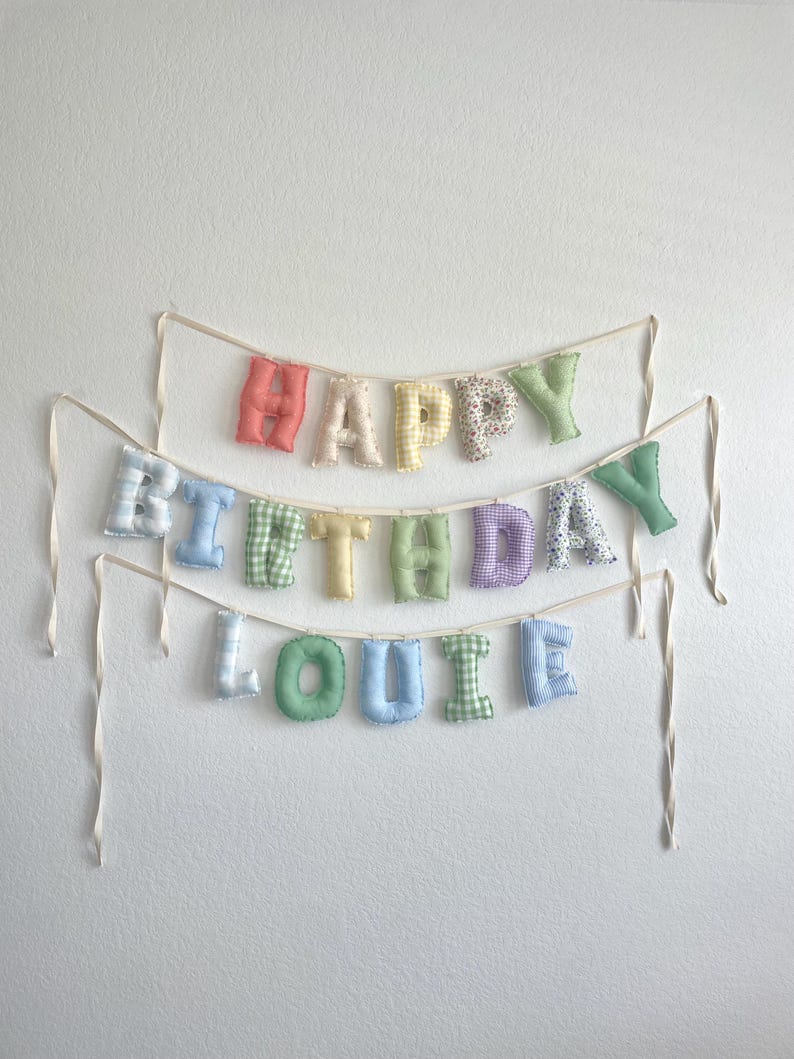 K&ouml;nnte beinhalten: Ein Stoffbanner mit ausgestopften, weichen Buchstaben, die HAPPY BIRTHDAY LOUIE buchstabieren. Die Buchstaben sind aus verschiedenen pastellfarbenen Stoffen mit Vichy-Karo-, Streifen- und Blumenmustern gefertigt und an d&uuml;nnen, cremefarbenen B&auml;ndern befestigt.