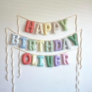 K&ouml;nnte beinhalten: Ein dreistufiges Stoffbanner mit ausgestopften, weichen Buchstaben, die HAPPY BIRTHDAY OLIVER buchstabieren. Jeder Buchstabe ist aus verschiedenen pastellfarbenen Stoffen mit Mustern wie Vichy-Karo, Blumen und Punkten gefertigt und an cremefarbenen B&auml;ndern befestigt.