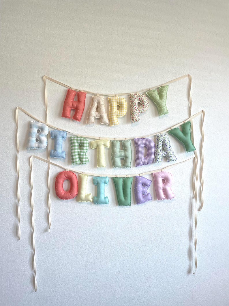 K&ouml;nnte beinhalten: Ein dreistufiges Stoffbanner mit ausgestopften, weichen Buchstaben, die HAPPY BIRTHDAY OLIVER buchstabieren. Jeder Buchstabe ist aus verschiedenen pastellfarbenen Stoffen mit Mustern wie Vichy-Karo, Blumen und Punkten gefertigt und an cremefarbenen B&auml;ndern befestigt.