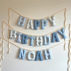 K&ouml;nnte beinhalten: Ein dreistufiges Stoffbanner mit ausgestopften, weichen Buchstaben in verschiedenen Blau- und Wei&szlig;t&ouml;nen. Die Buchstaben bilden den Schriftzug "HAPPY BIRTHDAY NOAH" und sind an d&uuml;nnen, cremefarbenen B&auml;ndern befestigt. Jeder Buchstabe hat ein Muster.